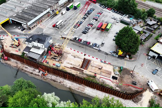 Aerial view of a riverside construction site with a large crane and a wall of tubular piles.