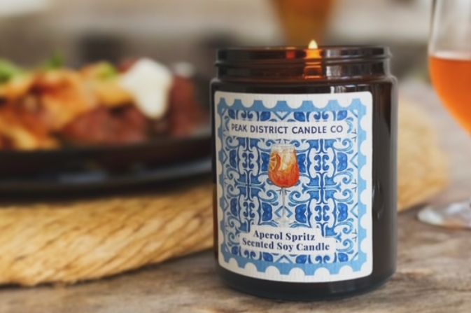 A close-up of a candle with an intricately designed blue label that reads Peak Districy Candle Co, Aperol Spritz scented soy candle