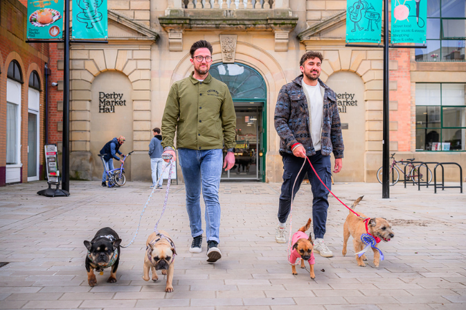 Two men each walking two small dogs on leads, with Derby Market Hall in the background