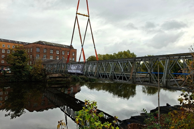 A large prefabricated bridge being craned over a river.