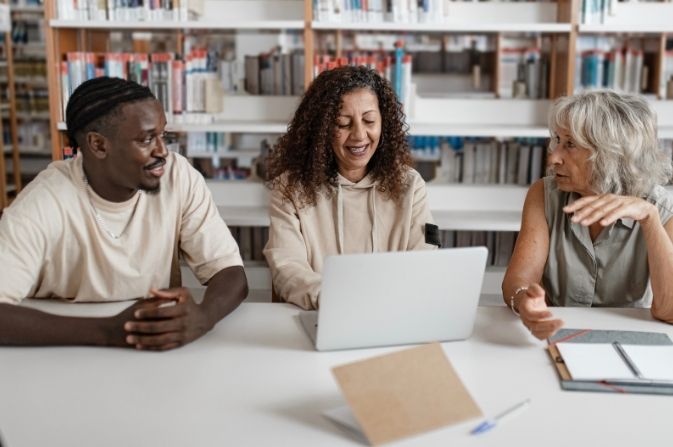 Three adults sit at a table in a library, talking. One is using a laptop computer.