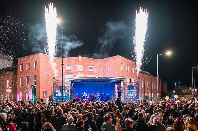 crowds of people in front of a stage as pyrotecnics light up the night sky