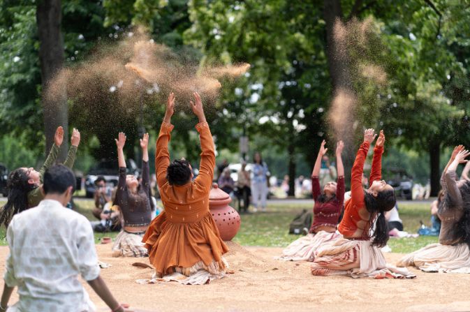 A group of kneeling performers in brightly coloured clothing throwing coloured dust into the air