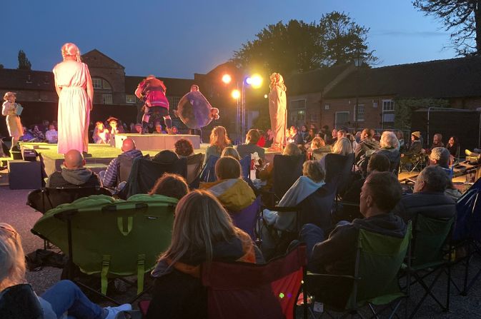 an audience sitting on camping chairs in a park watches actors giving a theatre performance as it goes dark