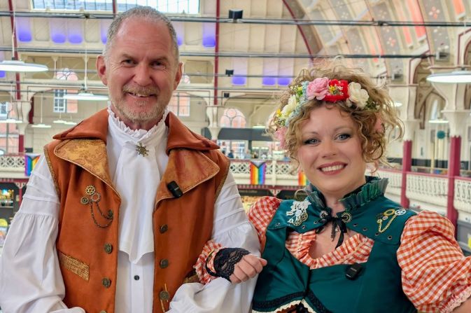A smiling man and woman dressed in traditional German clothing, standing inside Derby Market Hall