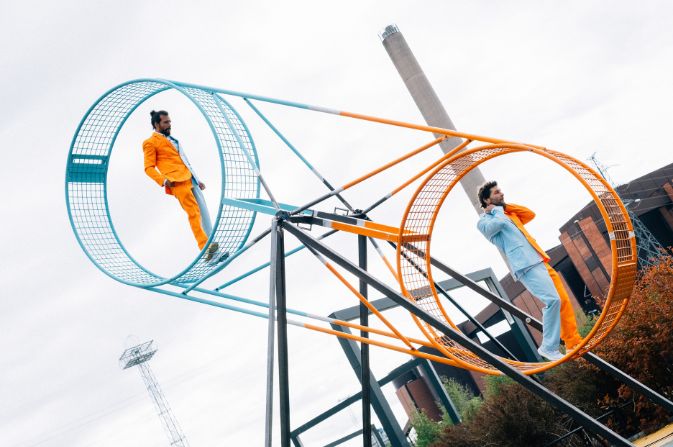 two male performers each balance inside circles at either end of a giant metal structure