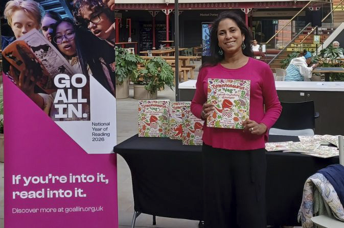 A smiling female author holding up a copy of her children's book, inside Derby Market Hall
