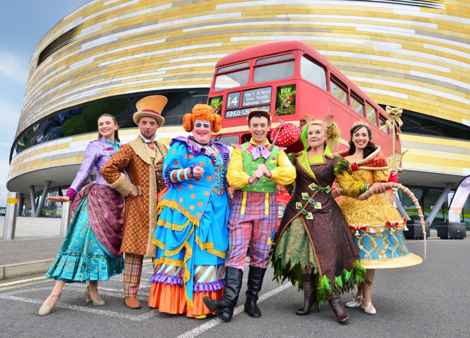 The colourful cast of Dick Whittington stand in front of a red double-decker bus, outside Derby Arena