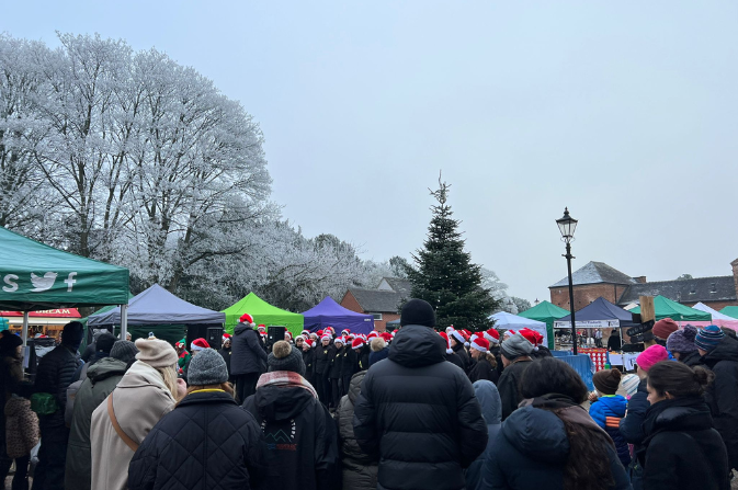 A large group of people gather to watch a festive choir sing in front of a Christmas tree