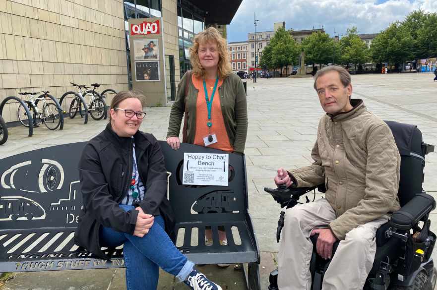 Beckie Hudson from the Council's Neighbourhoods team sits on a bench in the Market Place which has a sign to say it's a 'Happy to Chat' bench. Next to the bench is volunteer Rob Malcolm in his wheelchair, with Rachael Clark from the Neighbourhoods team standing behind the bench.