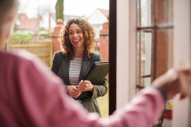 A smiling female social worker is welcomed in through the front door of a person's home