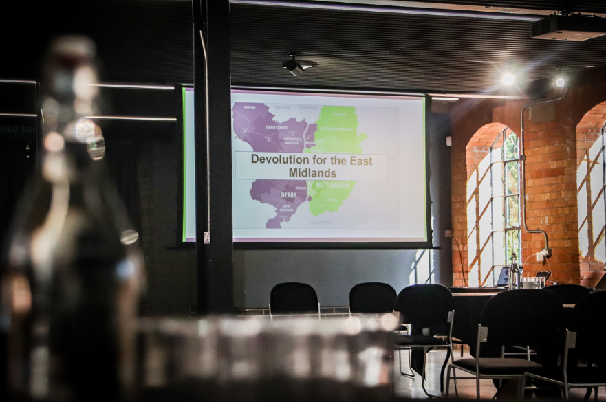 Large presentations screen displaying Devolution for the East Midlands. There is are rows of seats in front of the screen.