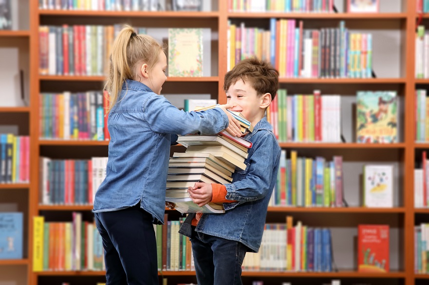 Two children carrying a stack of books in front of bookshelves