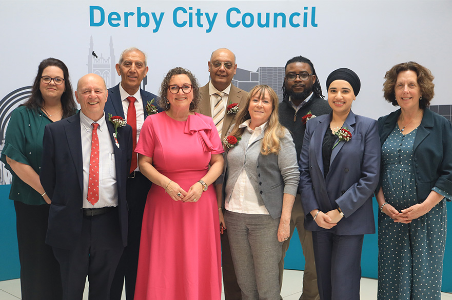 Group of people in professional dress looking at the camera. They are in front of a backdrop that reads Derby City Council.