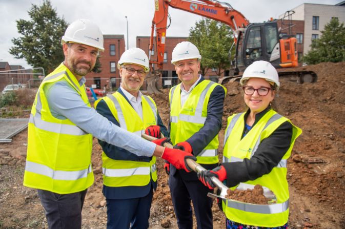 Four people in high vis jackets and hardhats. They are holding a spade and smiling at the camera.