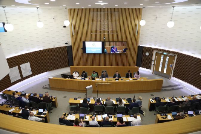 A large group of people gathered in a council chamber. They are having a meeting.