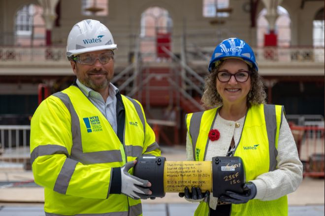 two people in high visibility jackets and hard hats holding a yellow and black time capsule.