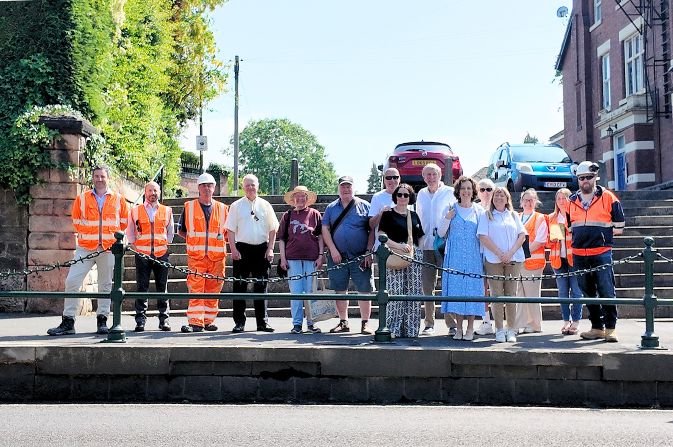 A group of people, some in high visibility outfits, stood on a raised pavement in front of a dark green metal railing with bollards and chains.