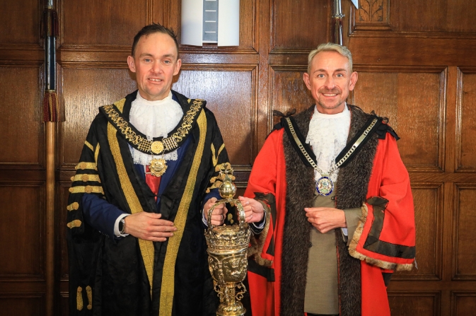 Two men in civic robes stood in front of a wood panelled wall and holding a gold mace.
