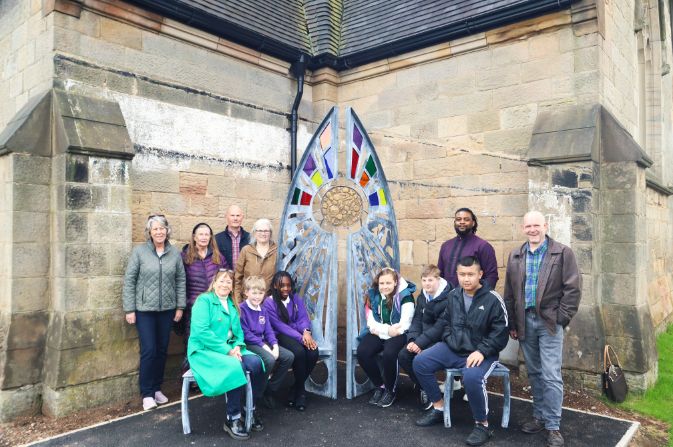 A group of people, a mixture of adults and school children, gathered around a large metal sculpture with stained glass elemnents.
