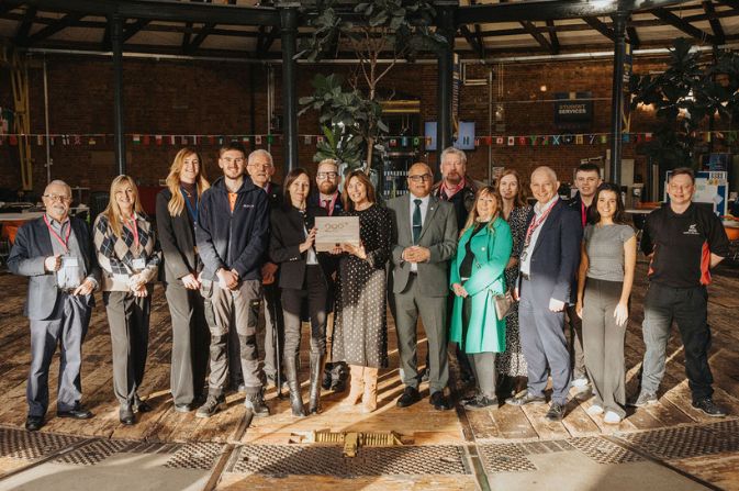 A large group of people stood for a group photo in a large, old industrial building. Two of them are holding a wooden box with the words Rail 200 engraved on the top.