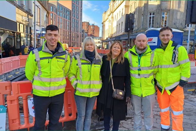 four people in high visibility jackets and a woman in a black coat stood in front of roadworks.