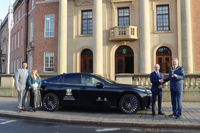 Four people stood around a black sedan car. It is a hydrogen powered Toyota Mirai.