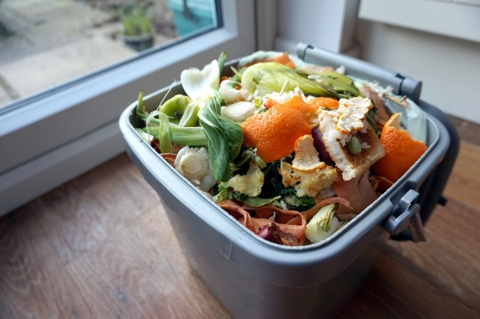 A grey kitchen caddy filled with mixed food waste such as fruit peels, vegetables, and leftovers, placed on a wooden floor near a window.